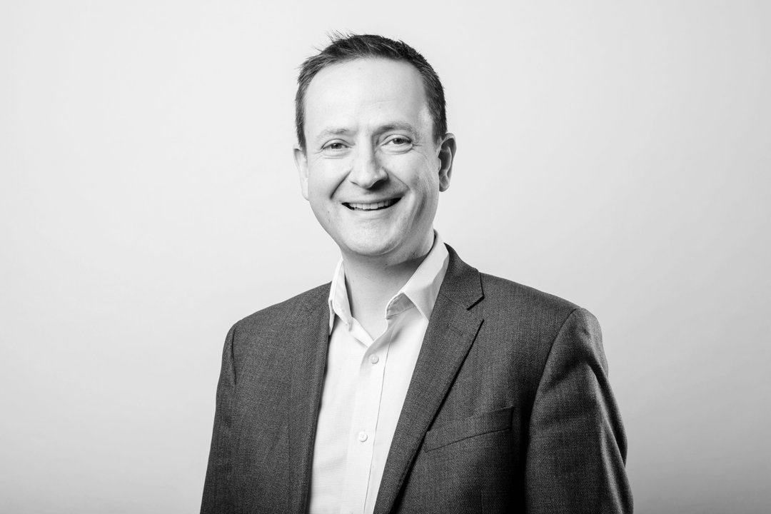 Smiling middle-aged man with short dark hair wearing a grey blazer and white dress shirt against a light studio background