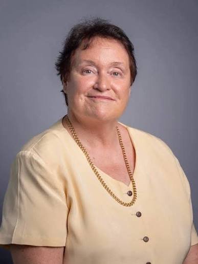 A middle-aged woman with short dark hair wearing a light yellow blouse and gold chain necklace, smiling softly against a gray
