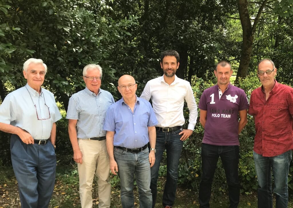 Six men of varying ages standing together outdoors in a lush green garden setting, wearing casual and business casual clothin