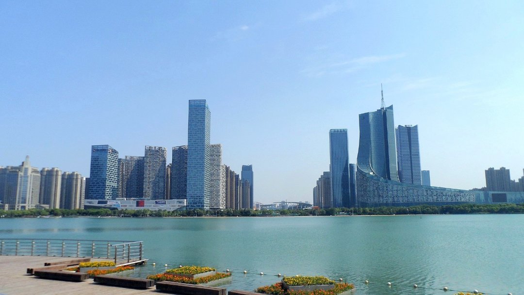 Modern glass skyscrapers of Shenyang Hunnan District rise above a calm green river with flowering waterfront promenade in for