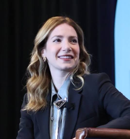 Smiling woman with wavy blonde hair wearing a black blazer and Prada triangle brooch seated at a conference panel