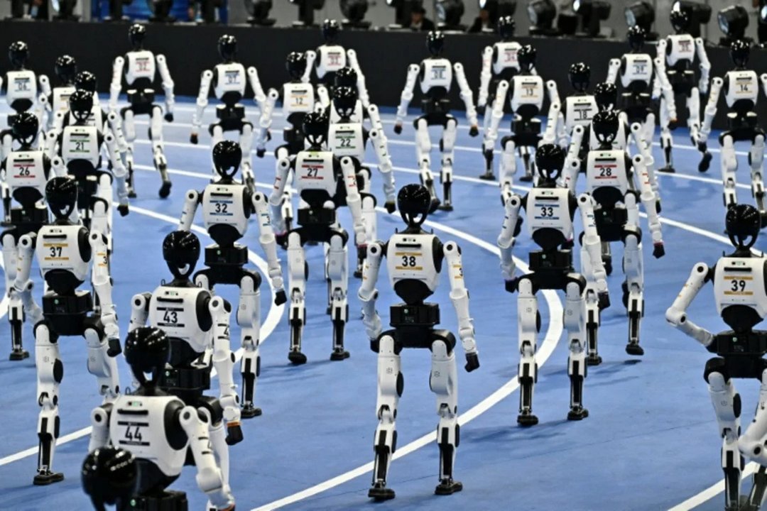 Dozens of white and black Unitree H1 bipedal humanoid robots with numbered race bibs walking on blue indoor track during robo