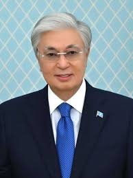 Older East Asian man in dark suit with blue tie and small flag lapel pin posed against light teal background in official port