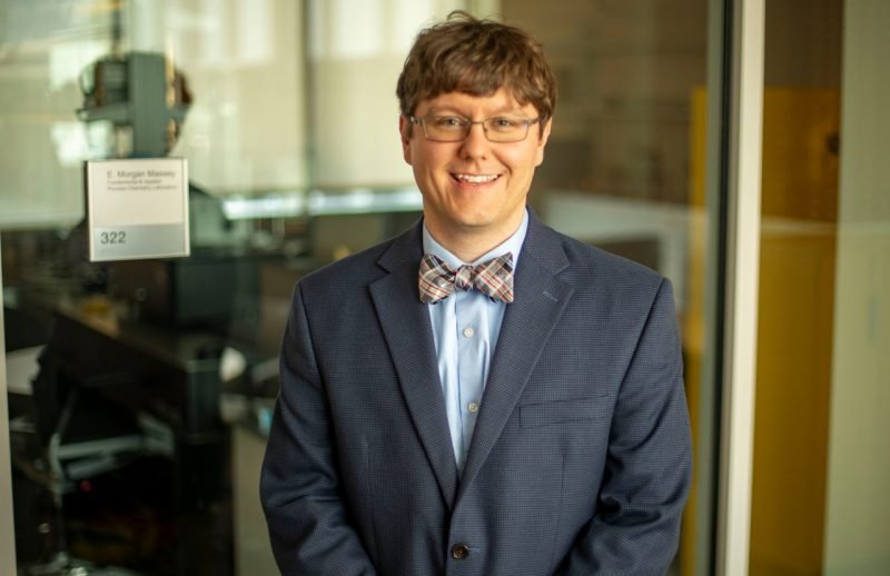 Smiling man in blue suit jacket, plaid bow tie, and glasses standing outside room 322 of the E. Morgan Massey laboratory