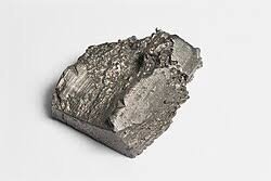A rough chunk of pure vanadium metal with silvery-gray crystalline surface texture and irregular jagged edges on white backgr