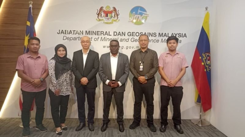 Six professionals posing at Jabatan Mineral Dan Geosains Malaysia headquarters in front of official signage with Malaysian fl