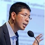 Young Asian man in dark suit speaking passionately at a podium with a microphone during a formal conference or seminar