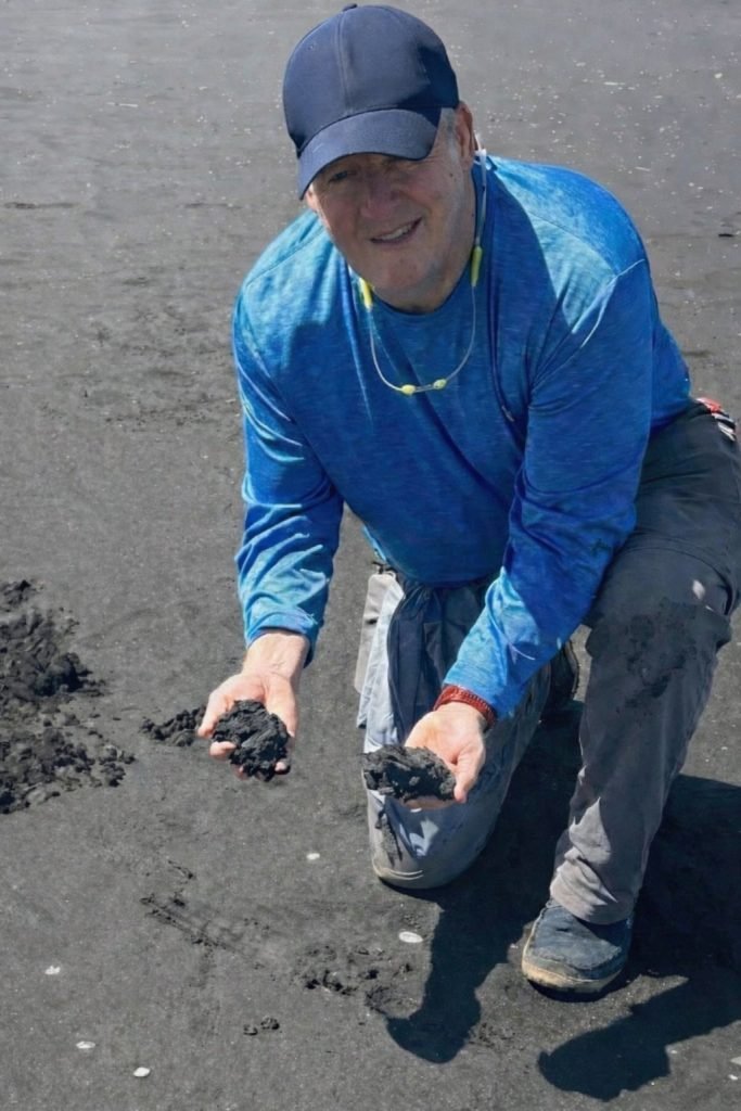 Smiling man in blue long-sleeve shirt and navy cap kneeling on black volcanic sand beach, holding handfuls of dark volcanic s