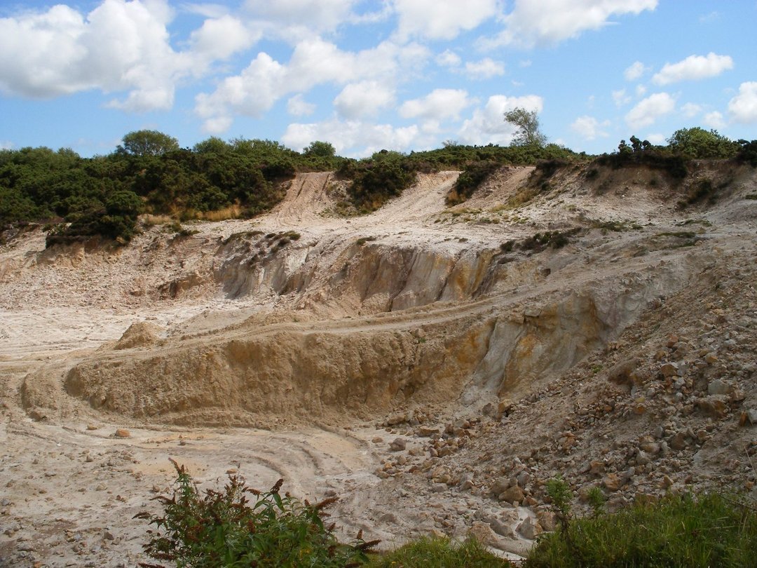 Open pit sand quarry with layered beige and grey sediment walls, rutted access tracks, green heathland shrubs along rim, blue