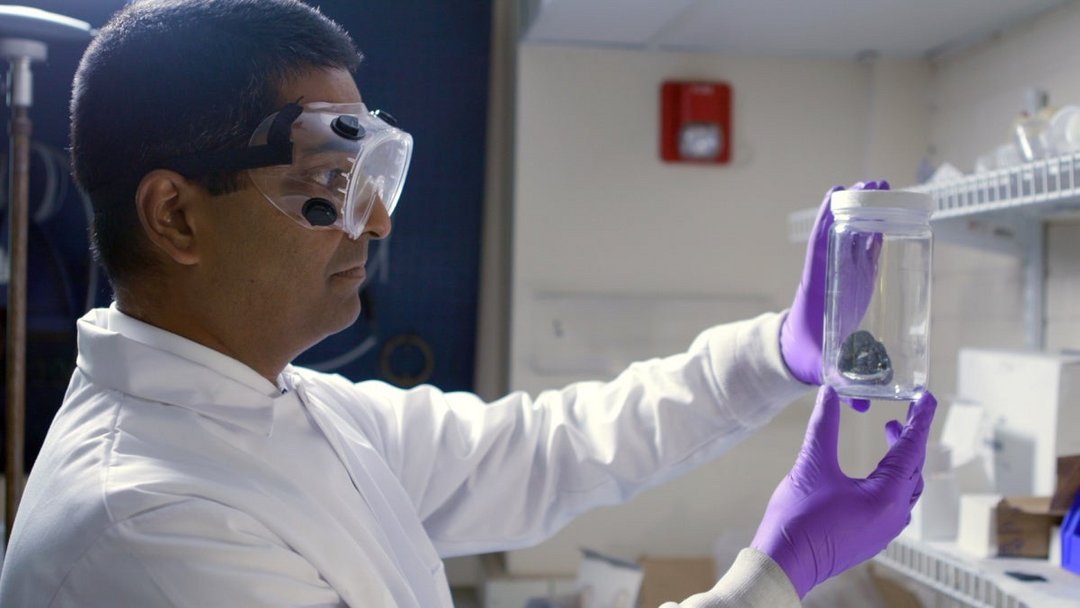 Male scientist in white lab coat and purple nitrile gloves holds clear glass jar with rock specimen up to light in research l