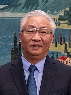 Senior Asian man with silver hair and glasses wearing a navy blue suit and teal tie standing before a landscape mural