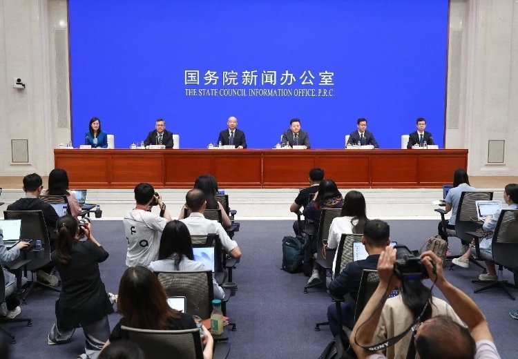 Six Chinese government officials seated at podium before blue State Council Information Office PRC banner with reporters and