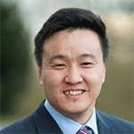 Smiling East Asian man in navy suit and striped tie with blurred outdoor greenery background, professional portrait
