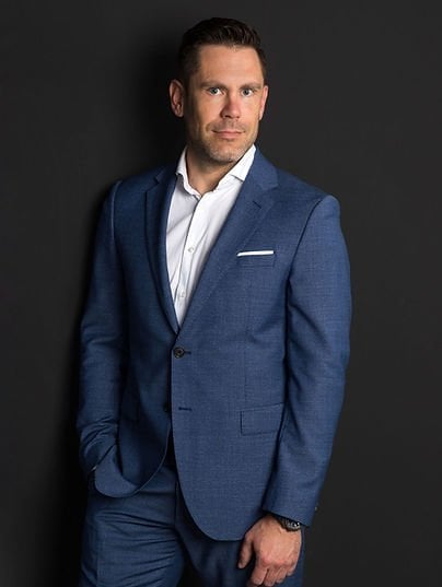 Man in tailored blue suit with white dress shirt and pocket square, leaning confidently against dark gray background