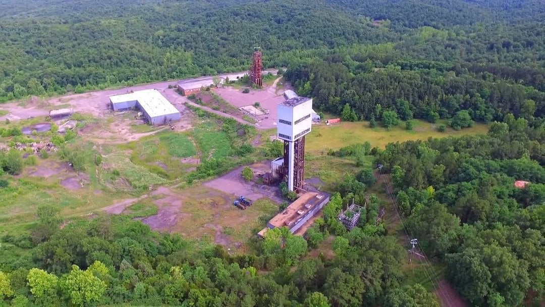 Aerial view of an abandoned coal mine with a rusted steel headframe tower, concrete shaft building, and derelict warehouses a