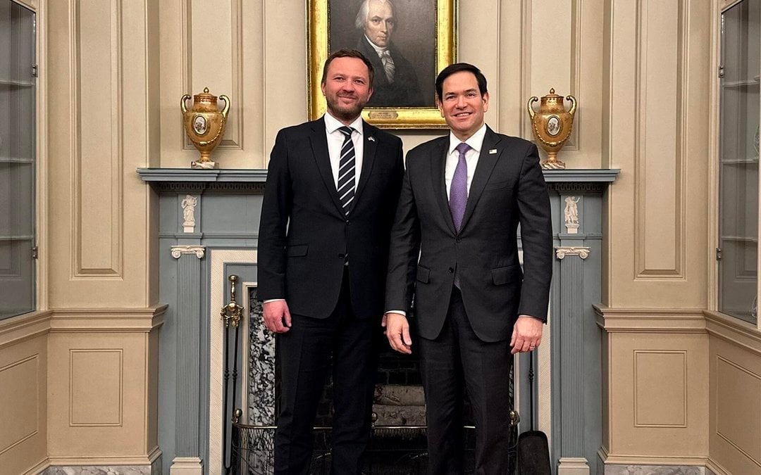 Two diplomats in dark suits standing before an ornate fireplace with golden urns and historical portrait in formal government