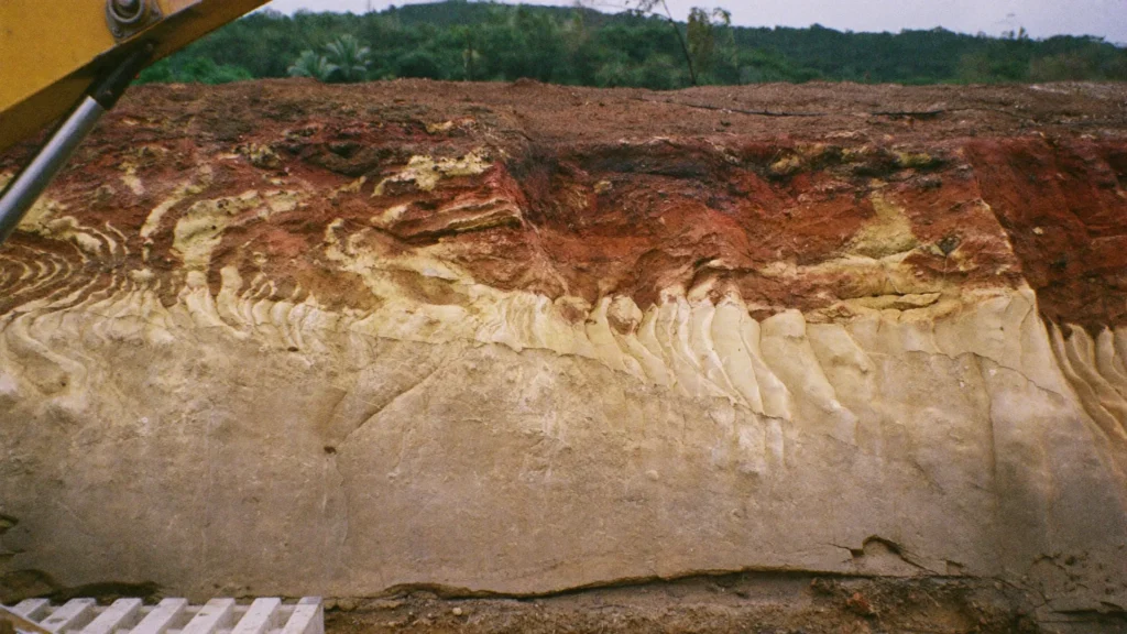 Freshly cut bench face of weathered ion-adsorption clay with pale and rust-red layers on a tropical hillside
