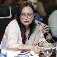 A woman wearing glasses and a pink blouse seated at a formal panel discussion table with a microphone in front of her