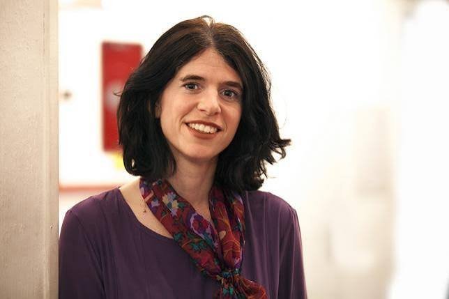 A smiling woman with short dark wavy hair wearing a purple blouse and colorful floral scarf, standing indoors near a white wa