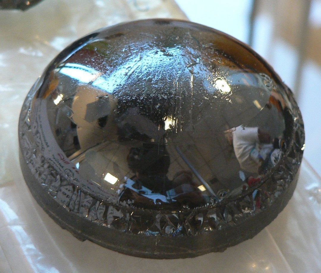 Highly polished convex Aztec obsidian scrying mirror with ornate carved base reflecting museum ceiling lights and visitors