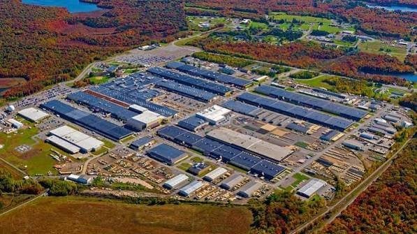 Vast industrial manufacturing campus with multiple warehouse buildings, parking lots, and autumn forest canopy in northeaster